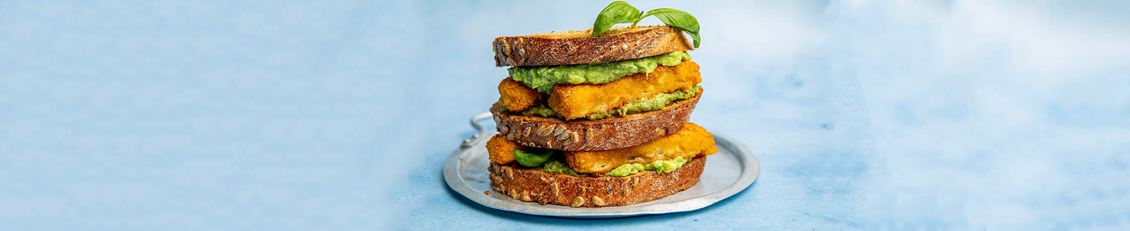 Sandwich med tre brødskiver med fisepinner og avokado på sølvfat med blåbakgrunn